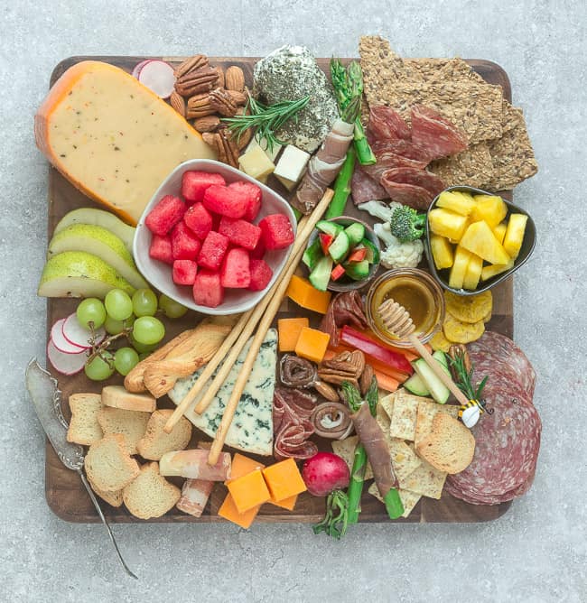 A cheese board full of meats, cheeses, and crackers, with a bowl of watermelon cubes and a bowl of pineapple cubes