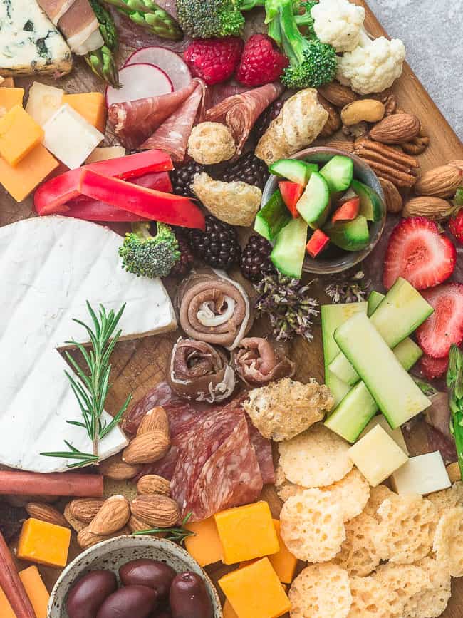 Close up of a cheese tray with a round of brie, a jar of cucumber slices, a bowl of olives, and lots of meat, cheese, and veggies