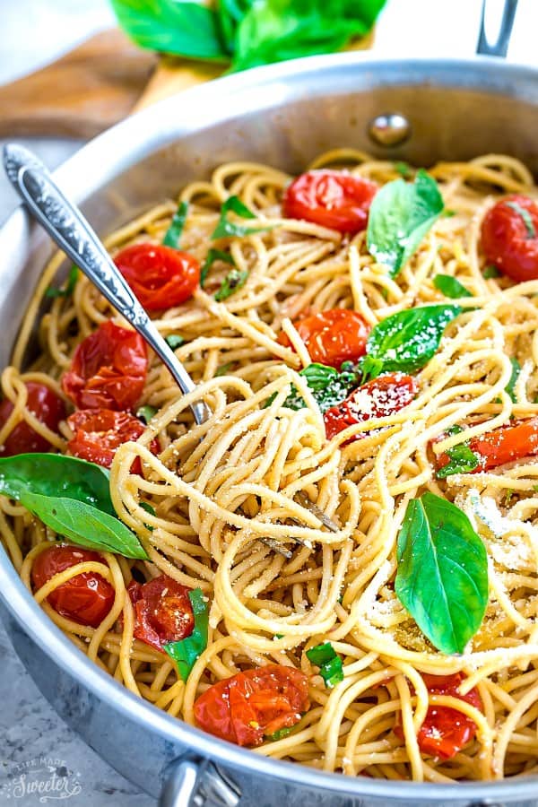 Close up of a bowl of cherry tomato pasta, topped with basil leaves and parmesan cheese, with a serving fork in the pan, with pasta wrapped around it. 