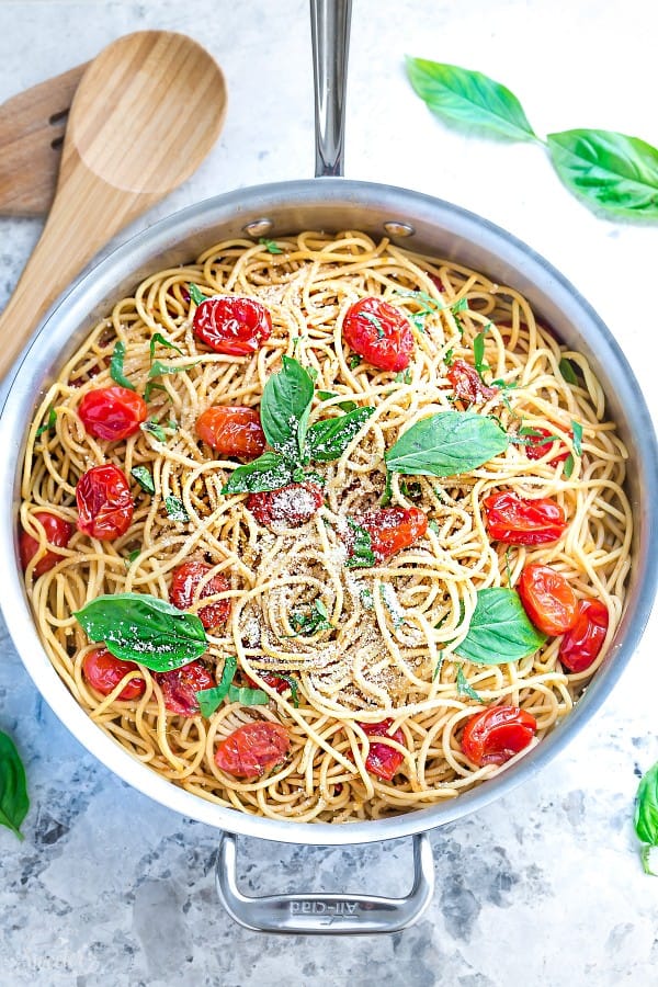 Overhead view of a pot of cherry tomato pasta, topped with whole basil leaves and parmesan cheese, next to wooden serving spoons and basil leaves.