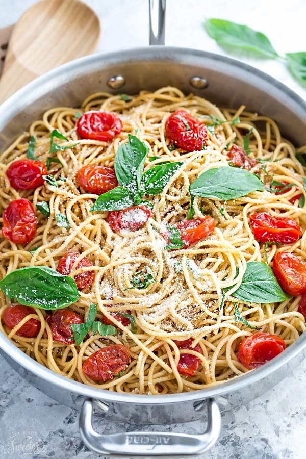 A pot filled with pasta, burst cherry tomatoes, and basil, topped with parmesan cheese and whole basil leaves, next to a wooden spoon.