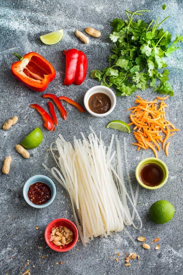 Grain free Chicken Pad Thai noodles, red bell peppers, peanutes, sliced carrots, crushed pepper and lime on a grey table.