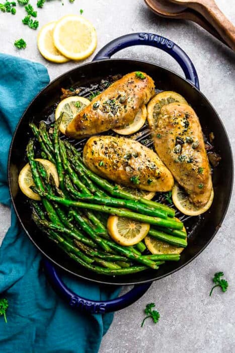 Top view of Easy Chicken Piccata with asparagus and capers in a blue cast iron pan on a grey background