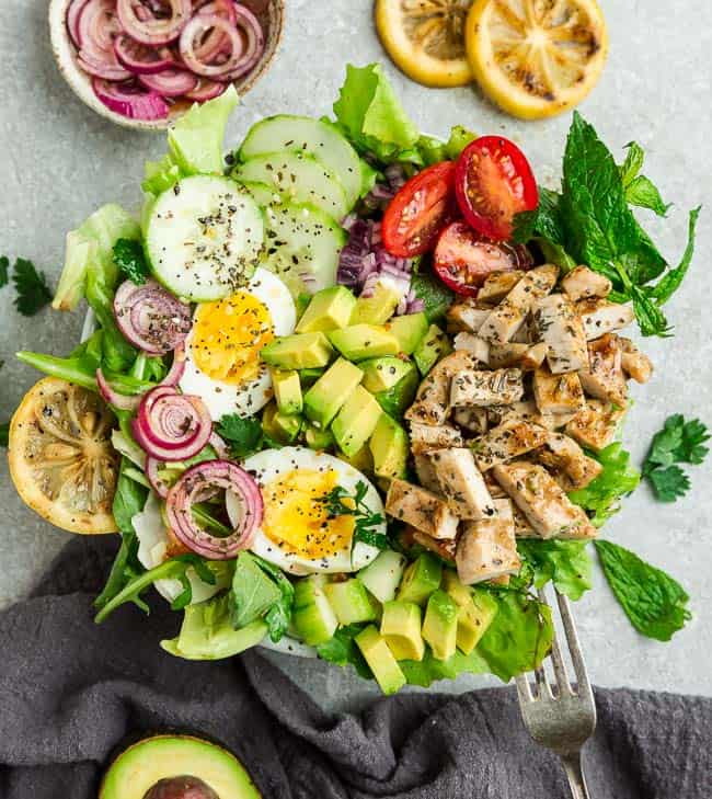 Top view of Chicken Shawarma Salad on a white bowl with avocado, hard boiled eggs with a fork on a grey background with grilled lemon slices