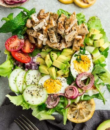 Top view of Chicken Shawarma Salad on a white bowl with avocado, hard boiled eggs with a fork on a grey background with grilled lemon slices