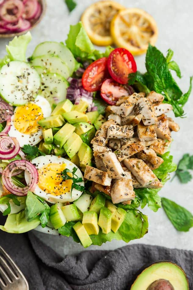 Top view of Chicken Shawarma Salad on a white bowl with avocado, hard boiled eggs with a fork on a grey background with grilled lemon slices