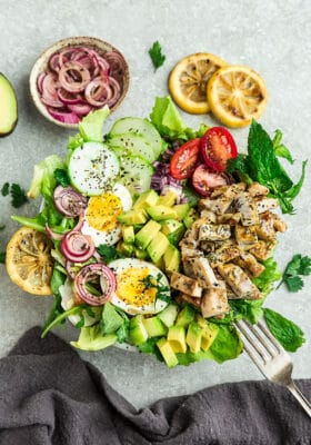 Top view of Chicken Shawarma Salad on a white bowl with avocado, hard boiled eggs with a fork on a grey background with grilled lemon slices