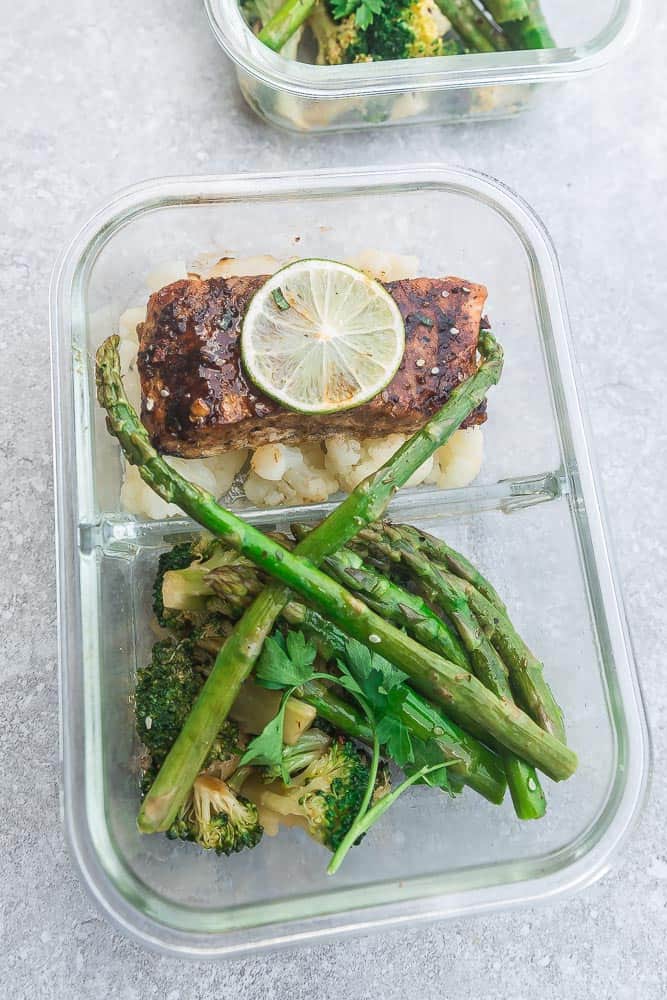 A divided leftover container with a salmon fillet topped with lime, asparagus, and broccoli