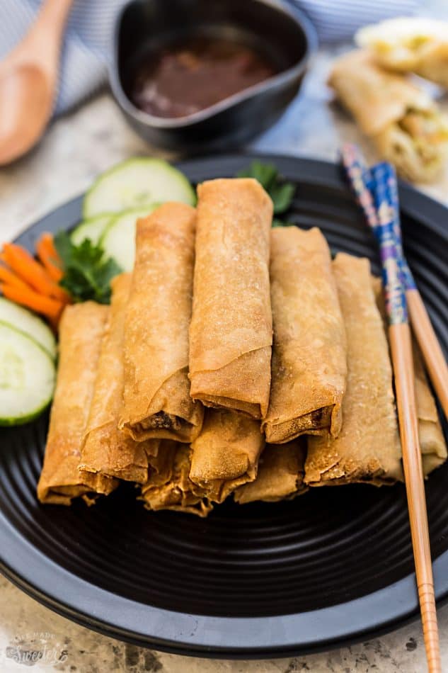 Chinese spring rolls piled onto a black plate with a dish of chili sauce behind it