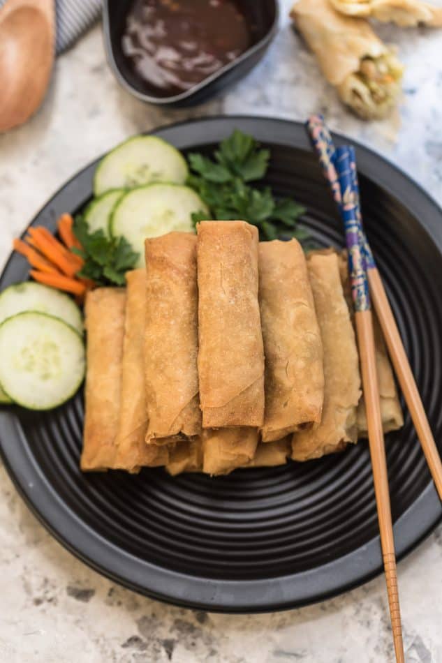 A plate full of Chinese spring rolls with a pair of wooden chopsticks beside them