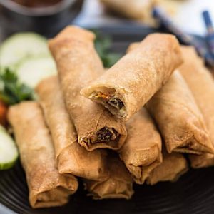 A stack of Chinese spring rolls on a round plate with veggies behind them