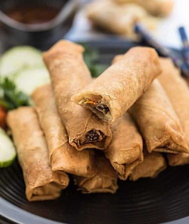 A stack of Chinese spring rolls on a round plate with veggies behind them