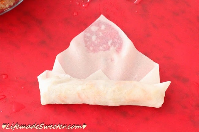 A Chinese egg roll in the process of being rolled on top of a bright red surface