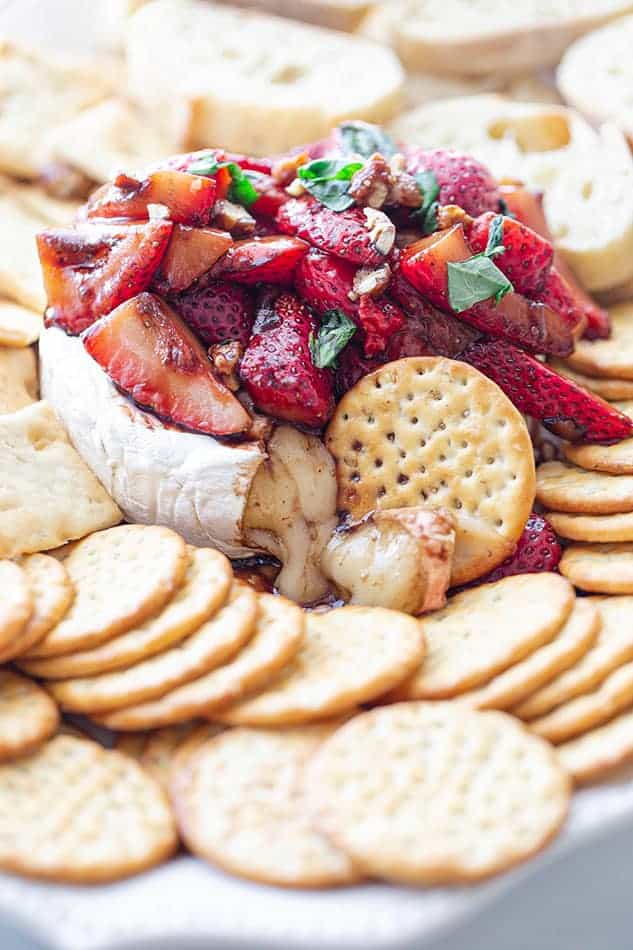 A pile of balsamic strawberries on top of a wheel of baked brie surrounded by gluten-free crackers and toasted baguettes
