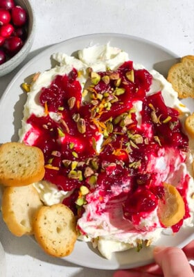 Whipped feta topped with homemade cranberry sauce and chopped pistachios in a white bowl surrounded by toasted baguettes.
