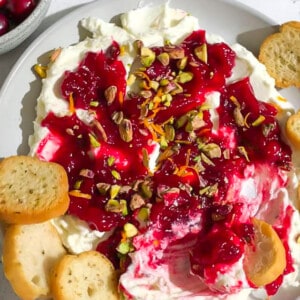 Whipped feta topped with homemade cranberry sauce and chopped pistachios in a white bowl surrounded by toasted baguettes.