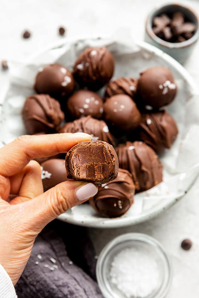 A hand holding a chocolate truffle with a bite out of it in front of a plate of chocolate truffles