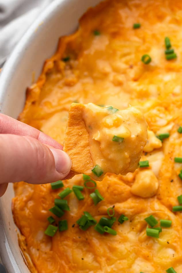 Close-up shot of a hand holding a tortilla chip over a batch of vegan buffalo chicken dip in a white round baking dish