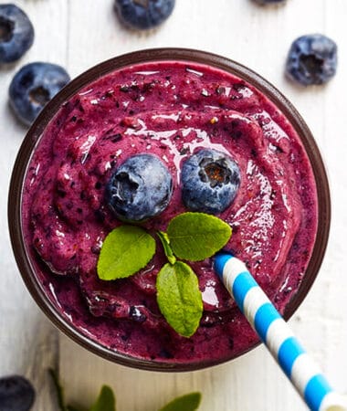 Top view of blueberry smoothie in a glass cup topped with fresh blueberries, mint and a blue striped straw