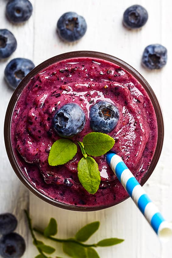 Top view of blueberry smoothie in a glass cup topped with fresh blueberries, mint and a blue striped straw