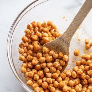 A batch of canned chickpeas with seasonings in a clear mixing bowl with a wooden spatula