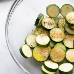 Zucchini slices inside of a large glass mixing bowl with oil, salt, pepper and garlic powder