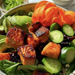 Seared salmon cubes over a bed of mixed salad greens, cucumber, ribboned carrots and avocado in a white bowl