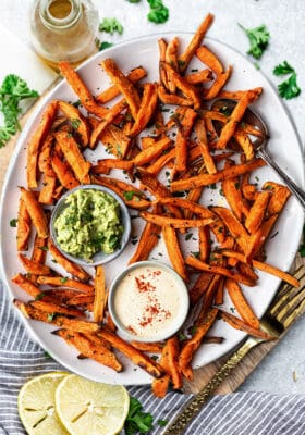 Top view of crispy sweet potato fries on a white oval plate with avocado dip and cashew dip