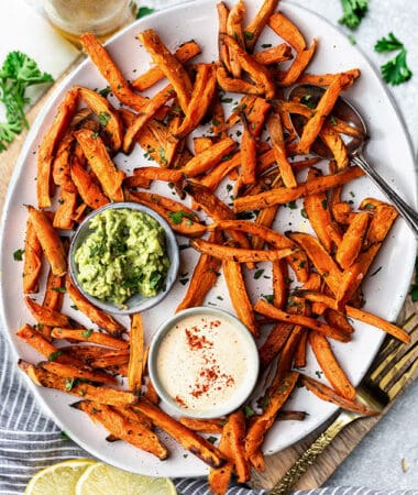 Top view of crispy sweet potato fries on a white oval plate with avocado dip and cashew dip