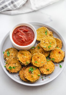 A pile of crispy zucchini chips on a white plate with a side of ketchup