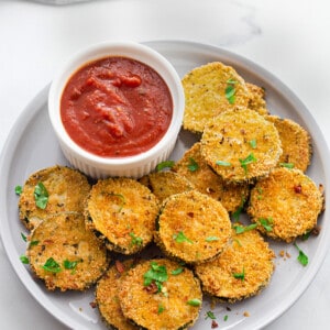 A pile of crispy zucchini chips on a white plate with a side of ketchup