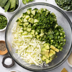 Overhead view of chopped green cabbage, kale, cucumbers, parsley, avocado and green onions side-by-side in a large clear mixing bowl to make a Green Cabbage Salad