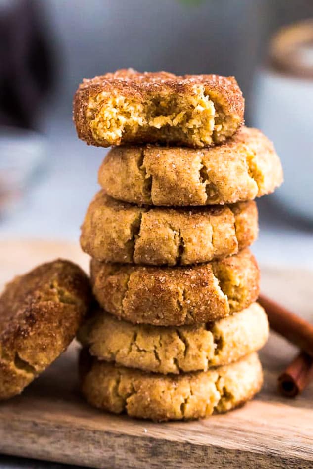 A stack of six snickerdoodle cookies, with a bite taken out of the top one, and a cookie rested against them, with cinnamon sticks in the background