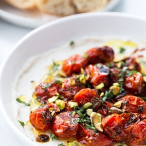 Side shot of roasted cherry tomatoes piled on whipped feta in a white bowl with a bowl of crusty bread on the side