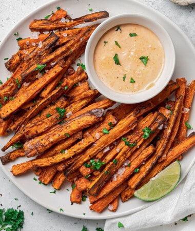 Close-up overhead shot of a pile of crispy air fryer carrot fries on a white plate with a side of dipping sauce and lemon