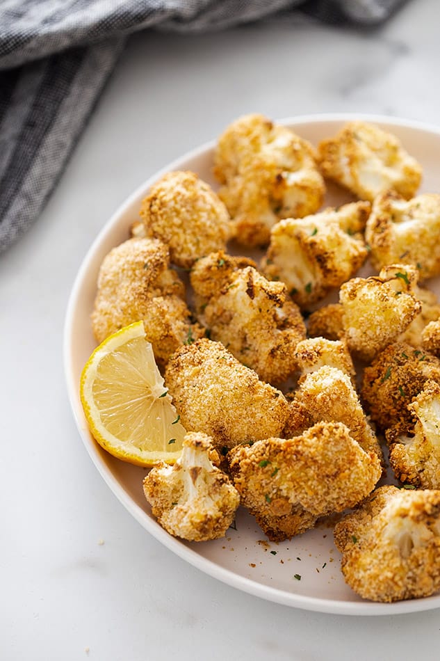 Crispy cauliflower wings piled onto a large plate with a lemon wedge beside them