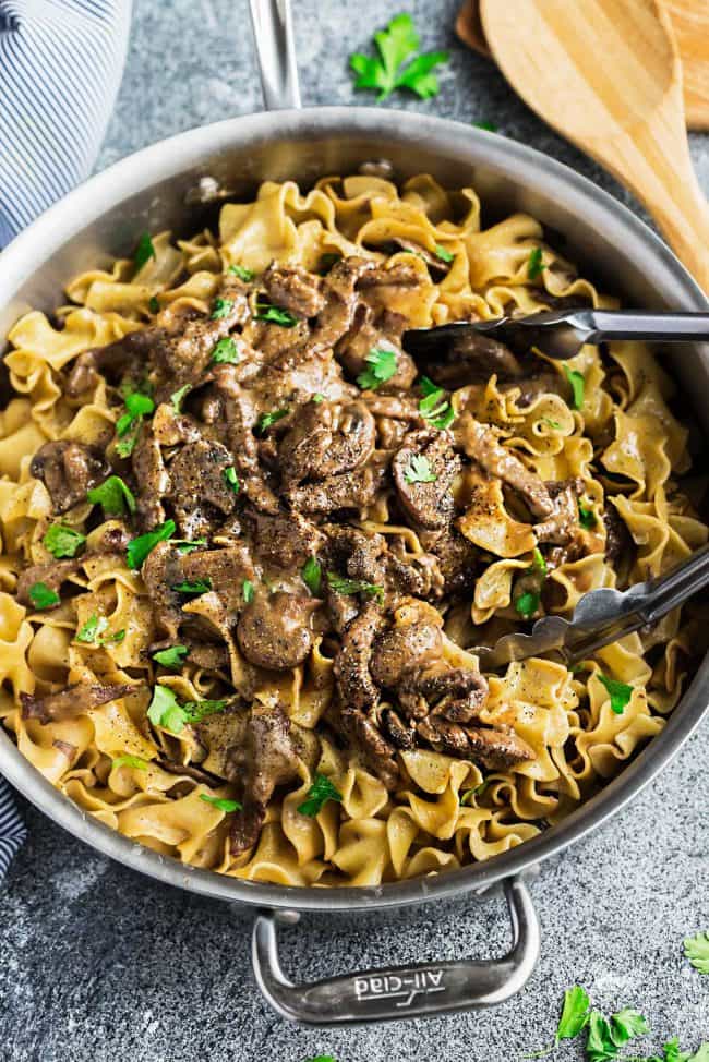 Delicious One Pot Beef Stroganoff being pulled out with a pair of silver tongs out of a cooking skillet.