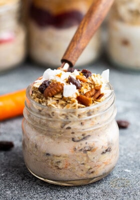 Side view of a jar of carrot cake overnight oats with chopped pecans and coconut on top with a spoon in the jar and a carrot in the background