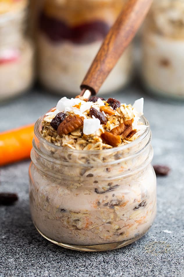 Side view of a jar of carrot cake overnight oats with chopped pecans and coconut on top with a spoon in the jar and a carrot in the background
