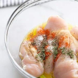 Raw chicken breasts in a clear bowl with seasonings