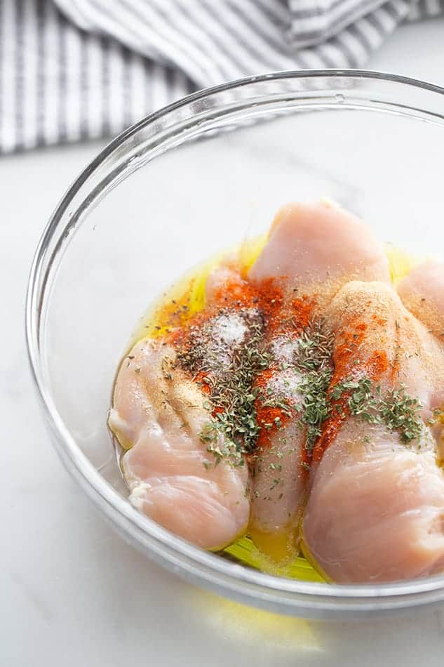 Raw chicken breasts in a clear bowl with seasonings