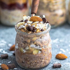 Side view of a jar of chocolate overnight oats with chopped chocolate chips, almonds and coconut on top with a spoon