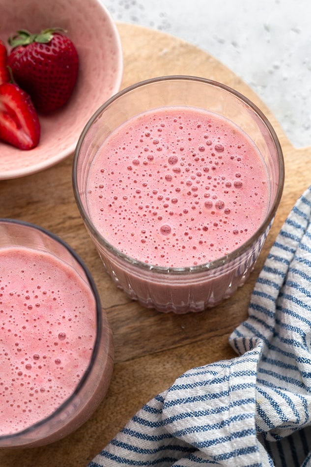 Two short glasses containing strawberry smoothies on a cutting board with a striped kitchen towel