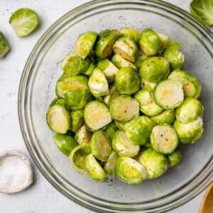 Top view of raw Brussels sprouts in a clear mixing bowl with oil and seasonings