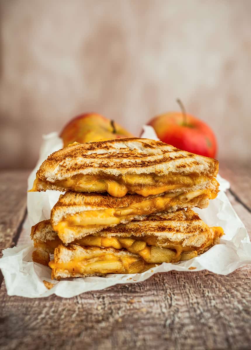 Four halves of grilled cheese sandwiches stacked on top of each other on parchment paper, with two apples behind them. 