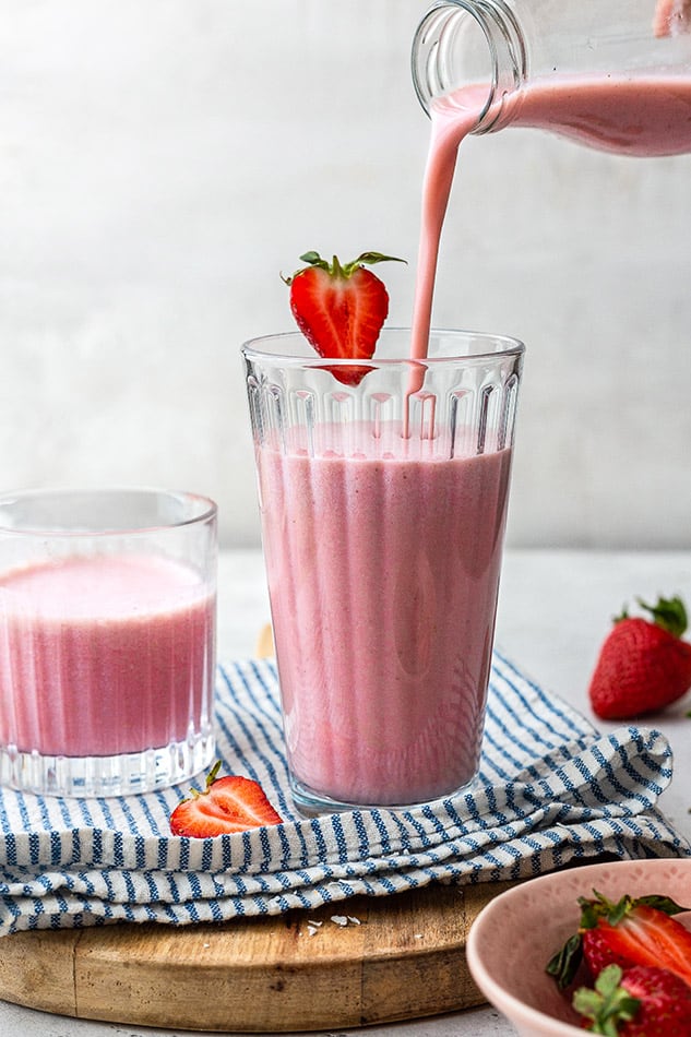A smoothie being poured from a glass bottle into a cup with a sliced strawberry on the rim