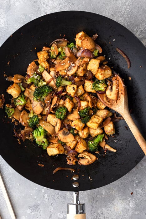 Top view of crispy cubed tofu, broccoli and mushrooms in a frying pan with a wooden spoon