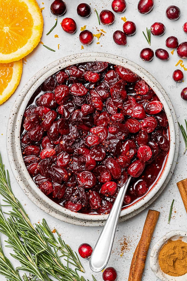 Overhead view of a bowl of homemade cranberry sauce with fresh herbs and spices nearby