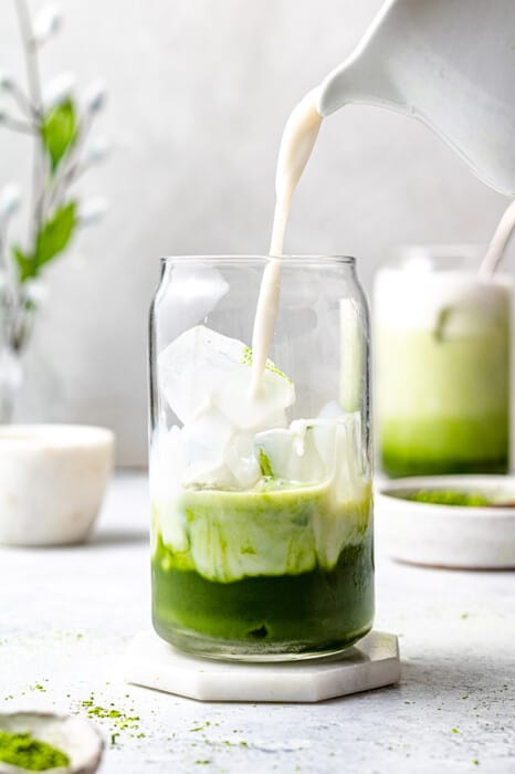 Side angle of one iced matcha in a glass and a jug of milk pouring milk into the glass