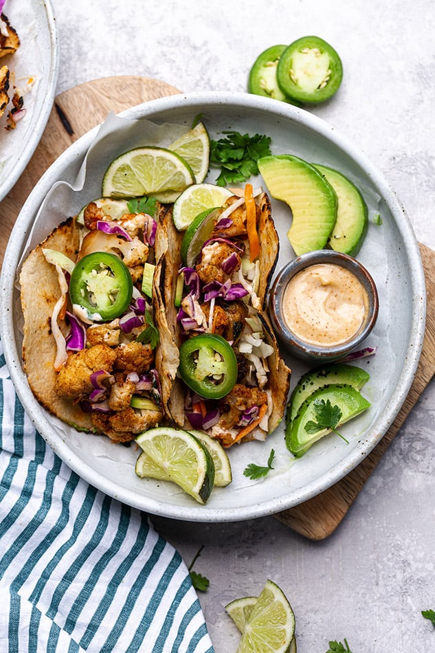 Overhead view of cauliflower tacos on a plate with limes, avocado, and jalapeno slices